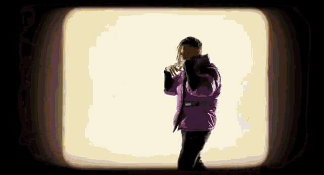 a man in a purple jacket is standing in front of a white wall and talking on a cell phone .