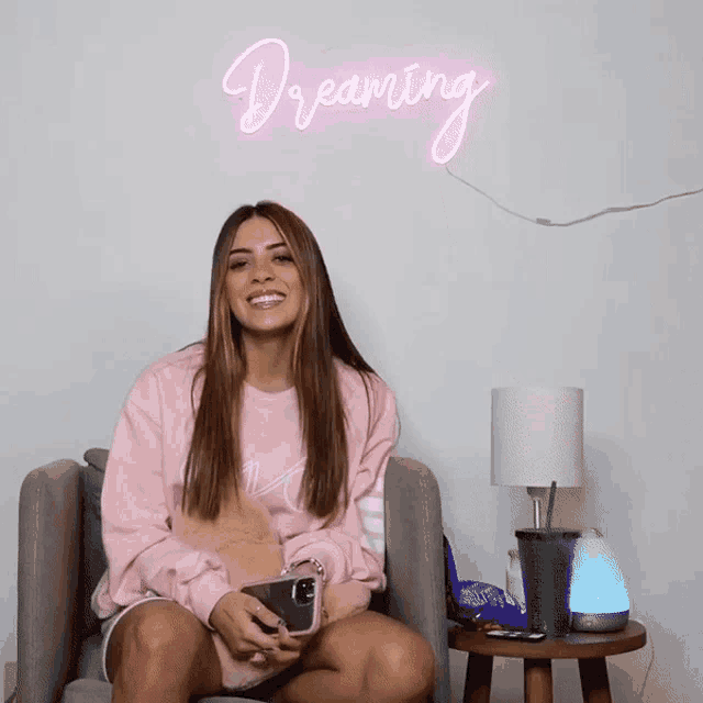 a woman in a pink sweatshirt is sitting in front of a neon sign that says dreaming