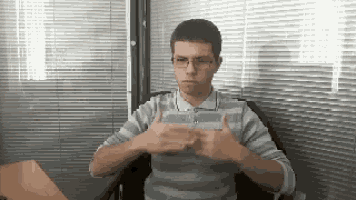 a man wearing glasses and a striped shirt is sitting in a chair and making a gesture with his hands .