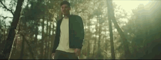 a man in a black jacket and white shirt is standing in a forest .