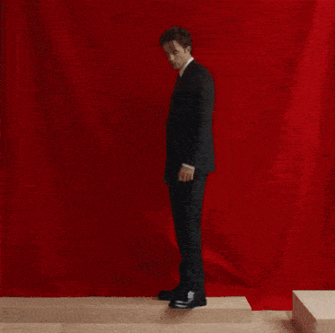 a man in a suit is leaning against a wall while another man stands in front of a red curtain