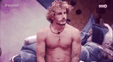 a shirtless man with curly blonde hair and a beard is sitting on a bed .