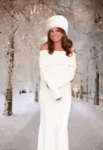 a woman in a white dress is standing in the snow wearing a white hat .