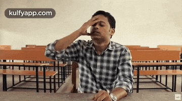 a man in a plaid shirt is sitting at a desk in a classroom with his hand on his face .