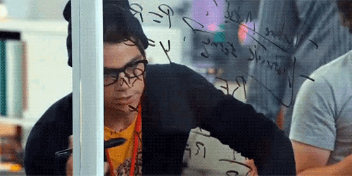 a man wearing glasses is writing on a glass wall with a marker .
