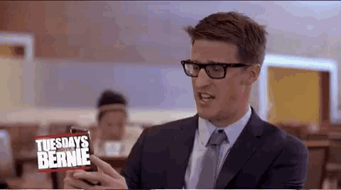 a man in a suit and tie is holding a cell phone in front of a sign that says tuesdays with bernie ..