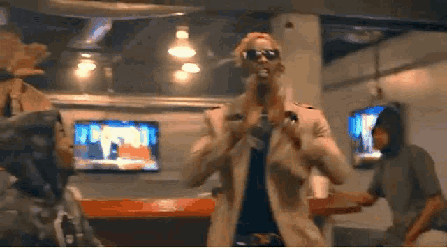 a man wearing sunglasses and a trench coat is singing into a microphone in a bar .