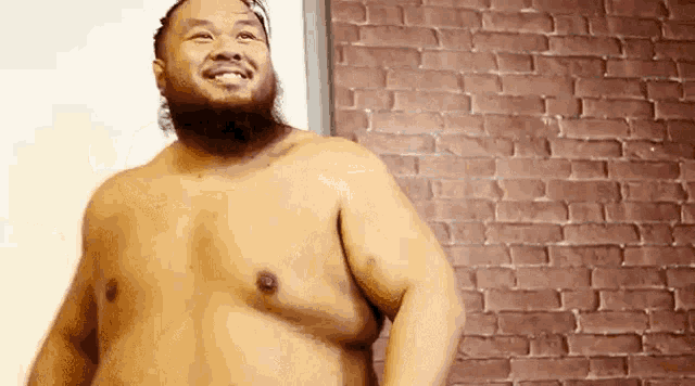 a shirtless man with a beard is smiling in front of a red brick wall .