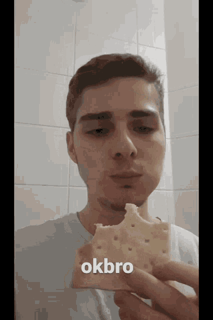 a man is holding a cracker with a bite taken out of it and says okbro on the bottom right