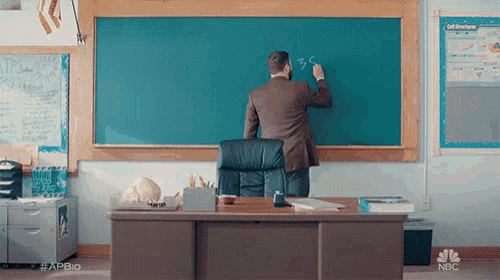 a man is writing on a blackboard in a classroom with nbc written on the desk