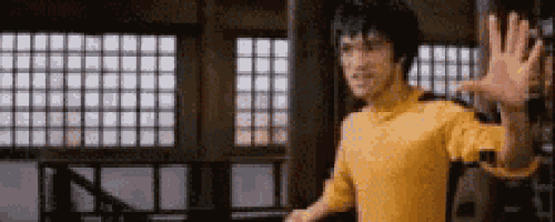 bruce lee is wearing a yellow shirt and waving his hand while standing in a room .