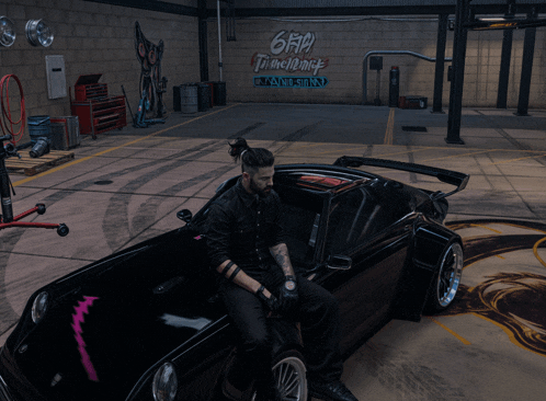 a man sits on the hood of a car in a garage with graffiti on the wall that says 666