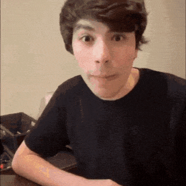 a young man in a black t-shirt is sitting at a table making a funny face .