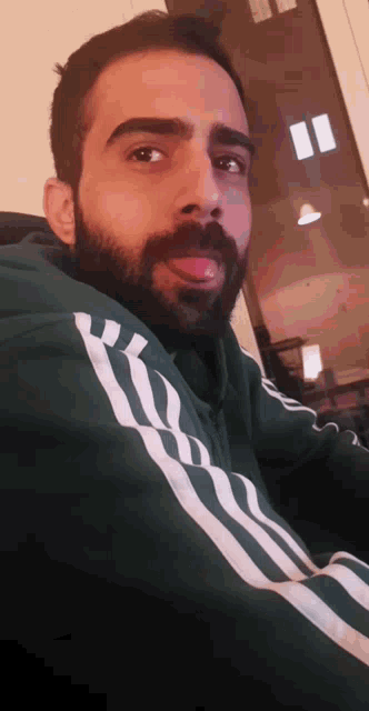 a man with a beard wearing a green adidas jacket sticks out his tongue
