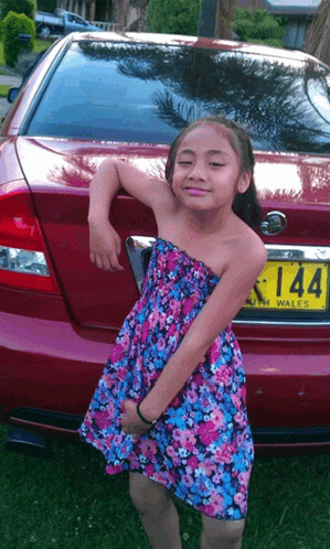 a girl in a floral dress is standing in front of a red car with a license plate that says 144
