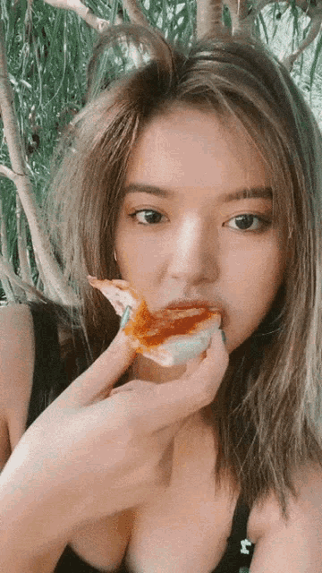 a woman in a black tank top is eating a slice of pizza with a tree in the background