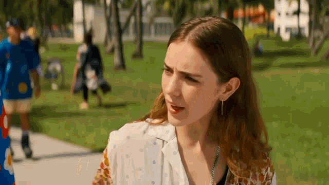 a woman is standing on a sidewalk in a park with a man in a blue shirt in the background .