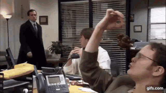 a man is sitting at a desk in an office taking a selfie with his phone .