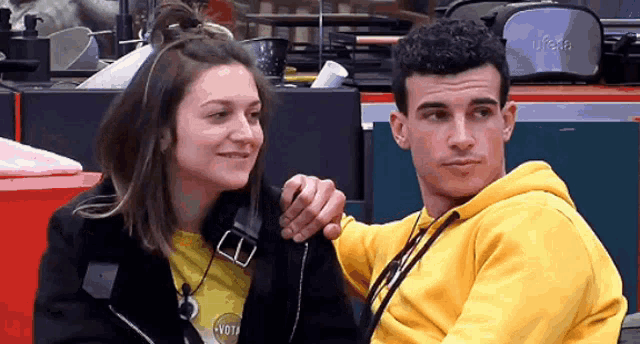 a man in a yellow hoodie holds a woman 's arm while they look at each other