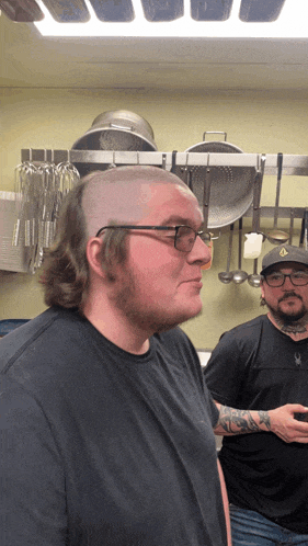 a man with glasses and a shaved head stands next to another man in a kitchen