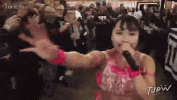 a woman singing into a microphone in front of a crowd with tjpw written on the bottom right