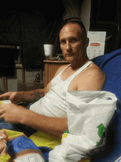 a man in a white tank top sits on a blue couch with a rubbermaid box in the background