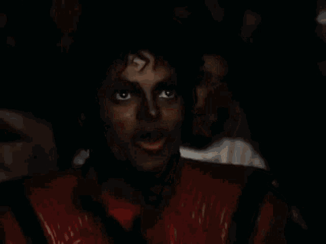 a man in a red leather jacket is sitting in a dark room with a crowd behind him .