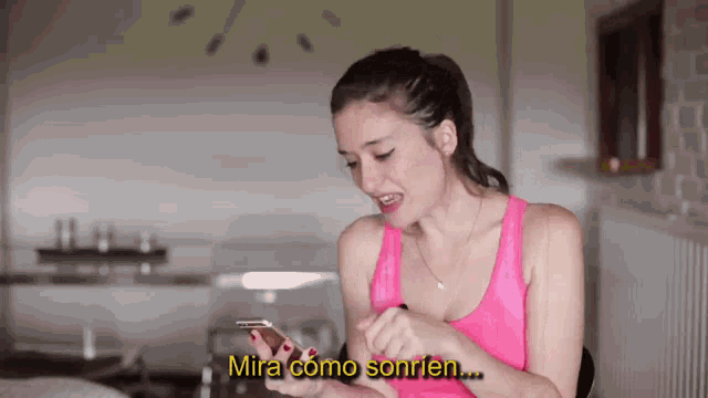 a woman in a pink tank top is holding a cell phone and says mira como sonrien