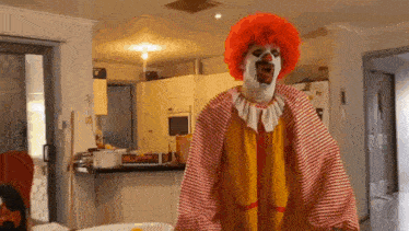 a clown in a red wig and yellow and red striped outfit is standing in a kitchen .