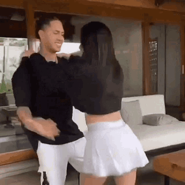 a man and a woman are dancing in a living room . the woman is wearing a white skirt and a black top .