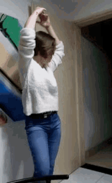 a woman in a white sweater and blue jeans is standing in a room with her hands in the air .