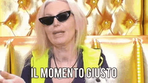a woman wearing sunglasses and a yellow vest is sitting on a couch and says il momento giusto .