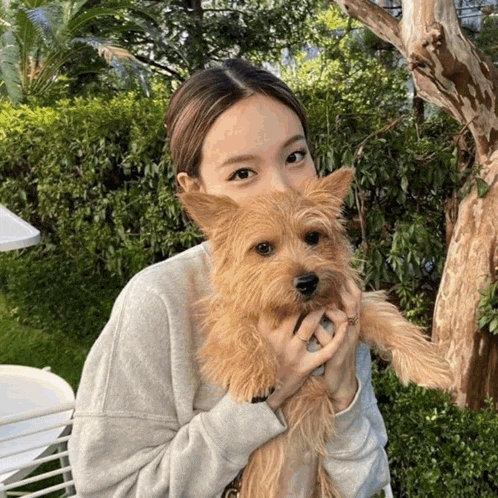 a woman holding a small brown dog with a tree in the background