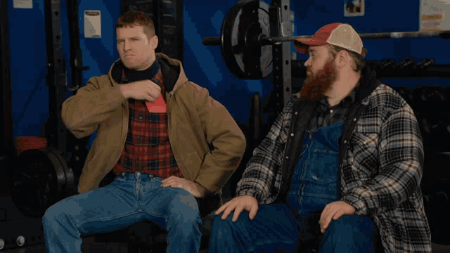 two men in plaid shirts and overalls sit in front of a barbell