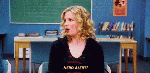 a woman is sitting in a chair in front of a chalkboard in a classroom and says `` nerd alert '' .