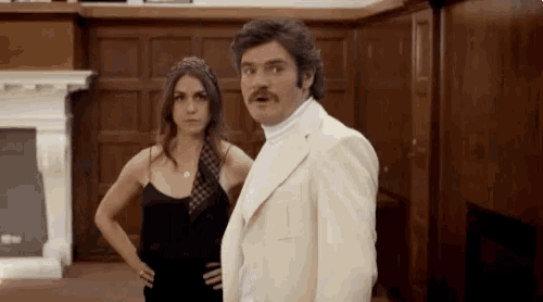 a man and a woman are standing next to each other in a room . the man is wearing a white suit and tie .