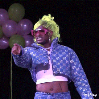 a man in a wig and sunglasses is pointing at something with balloons in the background