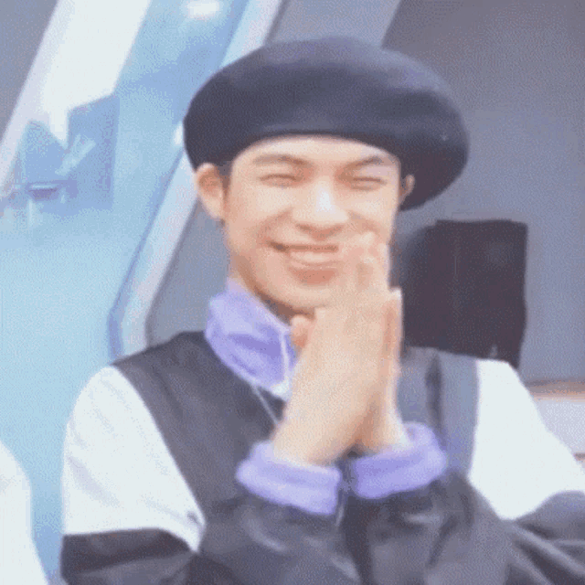 a young man wearing a black beret and a purple shirt is smiling with his hands together .