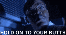a man with glasses is standing in a dark room with the words hold on to your butts above him .