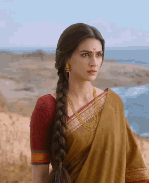 a woman wearing a braided ponytail and a necklace stands in front of the ocean