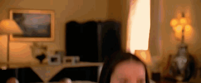 a woman is sitting on a bed in a living room with a lamp and a picture on the wall .