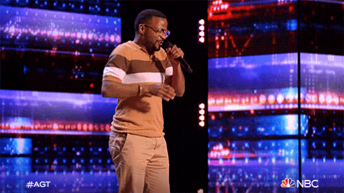 a man is singing into a microphone on a stage with a nbc logo in the background
