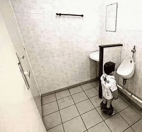 a little boy is urinating in a bathroom next to a urinal .