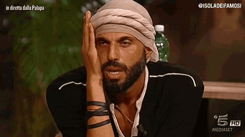 a man with a beard wearing a turban is sitting at a table with his hand on his forehead .