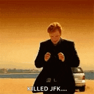 a man in a suit is standing on a beach with a car in the background and says `` killed jfk ... '' .