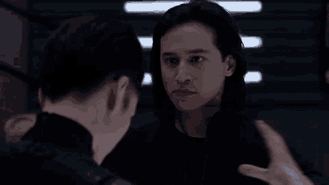 a man with long hair is talking to another man in a dark room .