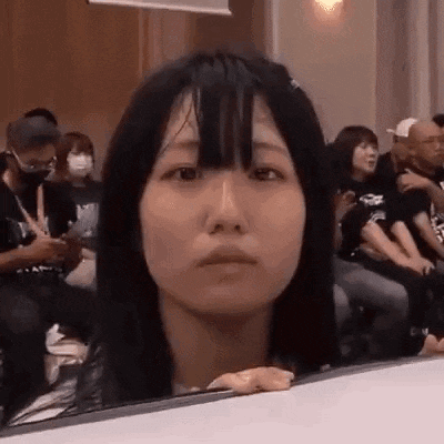 a woman with long black hair is sitting in a crowd of people and making a face .