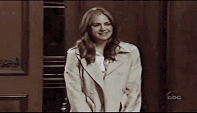 a woman in a trench coat stands in front of a door that says abc