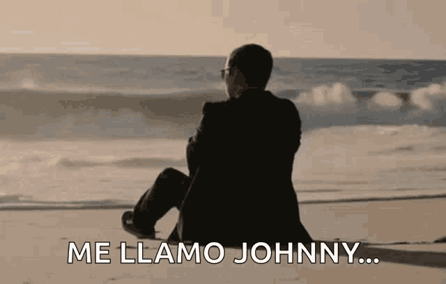 a man in a suit is sitting on the beach looking at the ocean and says `` me llama johnny '' .