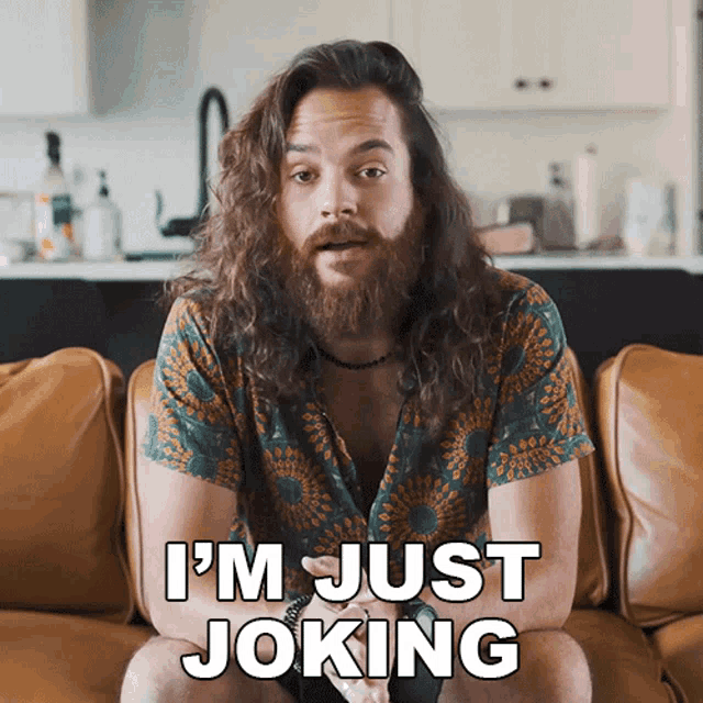 a man with long hair and a beard is sitting on a couch saying i 'm just joking